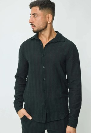 Homme aux cheveux courts et foncés et barbe, portant une chemise noire à manches longues boutonnée, regardant vers la gauche avec la main droite dans la poche, sur un fond uni.