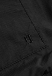 Svart tyg med en subtil textur, som visar en broderad logotyp i en mörkare nyans, placerad nära sömmen.