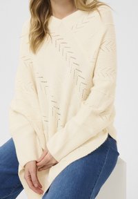 Krämfärgad stickad tröja med V-ringning, med texturerade fiskbensmönster och ribbade manschetter. Mjuk, avslappnad passform över blå jeans.