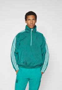 Blågrön pulloverjacka med hög krage, kvartsdragkedja, vita randiga detaljer och en framficka. Matchande blågröna byxor kompletterar looken.