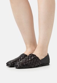 Des chaussures noires en cuir tressé à enfiler avec une semelle plate, présentant un motif tressé texturé et des accents en cuir lisse au niveau du talon.