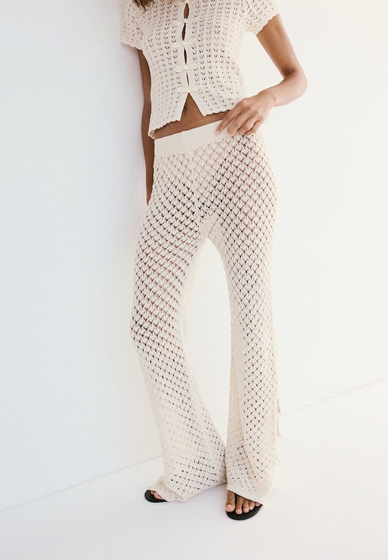Femme portant un haut en crochet crème à boutons et un pantalon large assorti, associé à des sandales noires à bout ouvert, debout contre un mur blanc.
