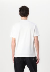 Witte katoenen t-shirt met een ronde hals en korte mouwen. Gladde textuur en een ontspannen pasvorm. Draagt samen met zwarte jeans met stiksels.