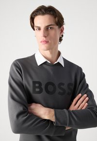 Γκρι φούτερ με μαύρο λογότυπο "BOSS", που διαθέτει στρογγυλή λαιμόκοψη και υφασμάτινα μανίκια πάνω από ένα λευκό πουκάμισο με γιακά. Λείος υλικός.