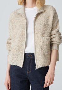 Beige textured zip-up jacket with a mock collar, ribbed cuffs, and a front pocket, paired with a white top and dark jeans.