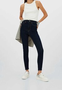 Mörkblå högmidjade jeans med orangea sömmar, kombinerade med en vit ribbad linne och vita sneakers med grå detaljer. Vertikala ränder på jackan.