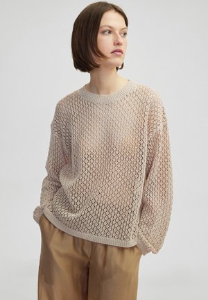 Pull en tricot beige clair avec un motif de tissage lâche, un large col rond et des coutures d'épaule tombantes, associé à un pantalon beige.