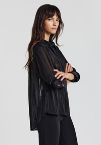 Frau mit langen braunen Haaren, die mit verschränkten Armen vor einem grauen Hintergrund steht. Sie trägt eine transparente schwarze gestreifte Bluse mit Knöpfen und schwarze Hosen.