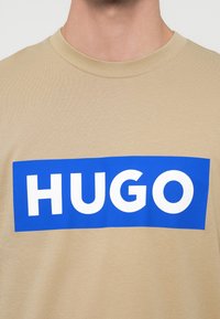Béžové tričko s okrúhlym výstrihom, na ktorom je na hrudi v strede výrazný modrý obdĺžnik s bielym textom "HUGO".
