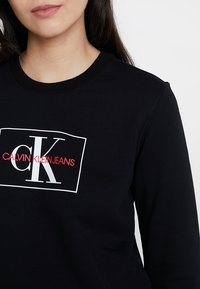 Kobieta w czarnej bluzie z białym prostokątnym logo, na którym widnieją duże litery "cK" oraz tekst "CALVIN KLEIN JEANS" w kolorze czerwonym.