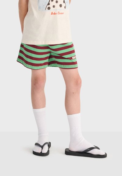 Enfant portant un short rayé vert et marron, des chaussettes blanches, des tongs noires et un t-shirt crème avec un motif imprimé.