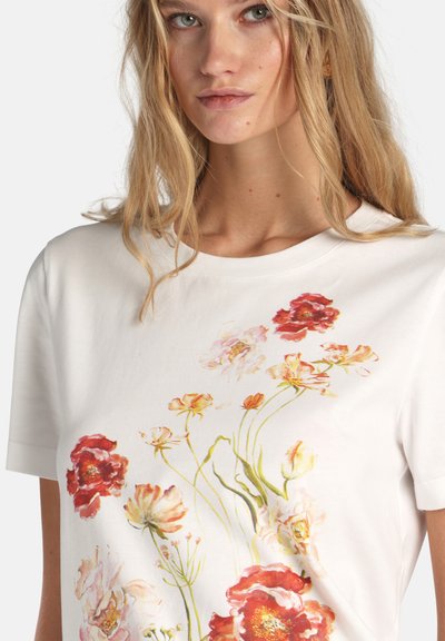 Blonde woman wearing a white T-shirt with a colorful floral print featuring red, yellow, and pink flowers and green stems.