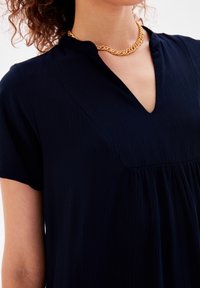 Kvinna som bär en strukturerad mörkblå V-ringad topp och ett tjockt guldvridet kedjehalsband mot en vit bakgrund.