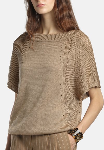 Femme portant un pull en maille beige à manches courtes avec un col large et un pantalon rayé marron, accessoirisée d'un bracelet manchette en écaille.