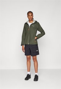 Grønn vanntett jakke med grå foring, glidelås og oransje detaljer. Kombinert med svarte shorts og svarte sportsko.