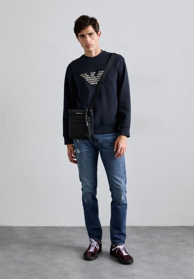 Sudadera azul marino con logo, bolso cruzado negro, jeans azules desgastados y zapatillas burdeos. Modelo de pie contra un fondo neutro.