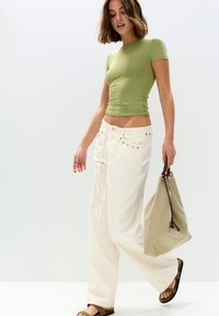Top scurt verde, ajustat, pantaloni albi cu talie înaltă și picioare largi cu detalii cu cuie, sandale brown tip slide, purtând o geantă de umăr verde deschis.