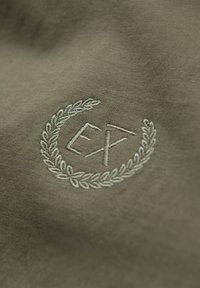 Primer plano de tela verde oliva con una corona de laurel blanca bordada y las letras "EX" en el centro.