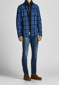 Niebieska koszula w kratę zapinana na guziki z dwoma kieszeniami na piersi, noszona na czarnej koszulce, w połączeniu z ciemnymi dżinsami i brązowymi sznurowanymi butami.