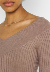 Maglione a coste marrone con ampio scollo a V, cuciture sulle spalle accentuate e una leggera lucentezza. Una collana in oro si posa sulla clavicola.