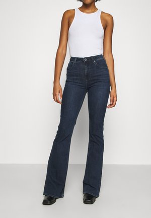 Vrouw draagt een witte mouwloze top, donkerblauwe hoge taille wijde jeans en zwarte schoenen, staand tegen een effen witte achtergrond.