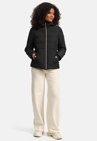 Black quilted puffer jacket with a high collar, a front zipper, and side pockets, worn over cream-colored pants.