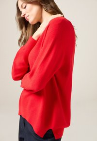 Czerwona sweter z długimi rękawami, miękką teksturą, luźnym krojem i zaokrąglonym dołem. Noszony na ciemnych dżinsach, zaprezentowany pod kątem.