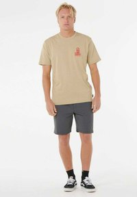 Camiseta de algodón beige con un logotipo gráfico rojo en el pecho, combinada con pantalones cortos gris oscuro. El calzado incluye zapatillas negras y blancas.