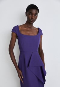 Lila Kleid mit kurzen Cap-Ärmeln, einem quadratischen Ausschnitt und einem asymmetrischen drapierten Detail am Saum. Glatte Stoffoberfläche.