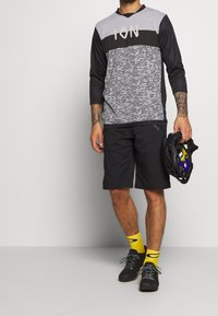 Musta ja harmaa urheilullinen pitkähihainen paita, jossa on camo-kuvio ja "ION"-logo, yhdistettynä mustiin shortseihin ja keltaisiin sukkuihin. Kypärä kädessä.