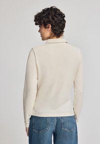 Långärmad krämfärgad polo skjorta med krage, med en slät textur och en kroppsnära passform, som matchas med medelhöga blå denimjeans.