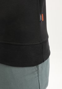 Zwarte katoenen sweatshirt met ribgebreide zoom, voorzien van een klein zijlabel in grijs en oranje. Gecombineerd met lichtgroene, geweven broeken.
