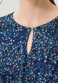 Bluse mit floralem Muster auf navy-blauem Grund, mit blauen und grünen Akzenten, einem Schlüsselloch-Ausschnitt und einem metallic Verschlussdetail am Hals.