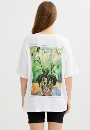 Mujer que lleva una camiseta blanca de gran tamaño con una pintura al óleo impresa de lirios blancos en un jarrón en la espalda.