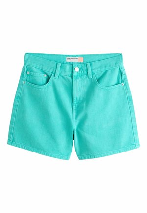 Giovane donna in piedi sulla spiaggia, indossa una camicia bianca con disegni di pesci blu e pantaloncini turchesi con le mani in tasca.