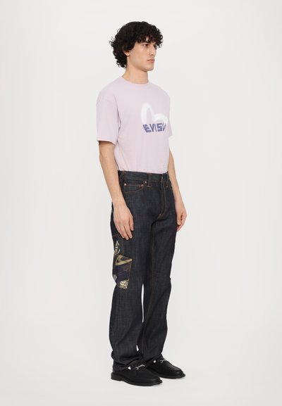 T-shirt violet clair avec logo, associé à un jean en denim sombre avec poches à motifs. Des chaussures noires complètent l'ensemble.