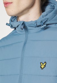 Svetle modrá bunda s vyp_paddingem, zapínáním na zip a nastavitelnou kapucí. Na hrudi se nachází černožluté logo orla.