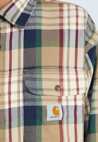 Camisa de cuadros en tonos beige, azul marino, verde y rojo. Presenta un bolsillo en el pecho y una etiqueta de marca con un logo amarillo. Tejido texturizado.