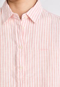 Pink and white striped linen shirt with a button-down collar, featuring beige buttons and subtle embroidered logo on the left side.