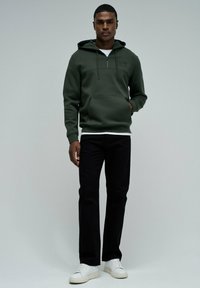 Sudadera verde oliva con bolsillo frontal, capucha con cordón y costuras visibles. Combinada con pantalones negros y zapatillas blancas.