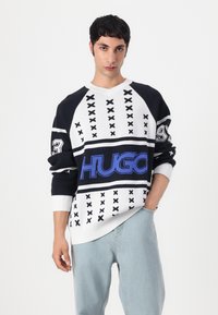 Svart-hvitt mønstrete genser med blå "HUGO" logo, V-hals design og ribbestrikkede mansjetter, kombinert med avslappede lyseblå jeans.