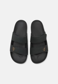 Mules noires avec deux sangles en tissu ajustables, une semelle intérieure texturée et un petit détail de logo marron. Design léger et doux.