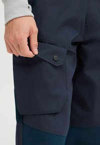 Navy cargo pants with a side pocket, featuring black snap buttons and a smooth texture. The design includes a tapered fit and multi-panel construction.