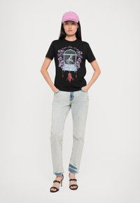 Black t-shirt featuring a colorful graphic print of a birdcage with floral accents. Paired with light blue jeans and black heeled sandals.