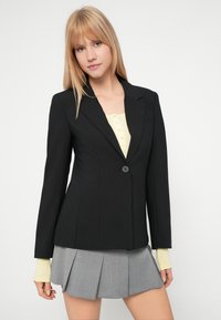 Μαύρο κομμένο σακάκι με κλείσιμο με μονό κουμπί, με πιεσμένα πέτα. Συνδυασμένο με ένα ανοιχτό κίτρινο τοπ και γκρι πλισέ φούστα.