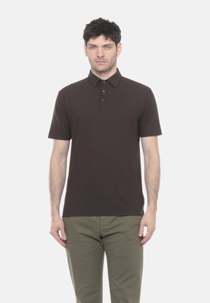 Uomo che indossa una polo a maniche corte marrone scuro e pantaloni verde oliva, in piedi contro uno sfondo bianco semplice.