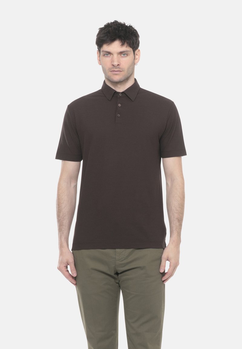 Uomo che indossa una polo a maniche corte marrone scuro e pantaloni verde oliva, in piedi contro uno sfondo bianco semplice.