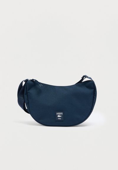 Sac bandoulière bleu marine en tissu texturé, présentant une forme arrondie, une sangle ajustable et une petite étiquette avec le logo de la marque à l'avant.
