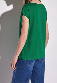 Woman wearing a green short-sleeve top and blue denim jeans, standing with her back to the camera against a plain background.