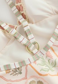 Bretelles réglables à carreaux avec anneaux dorés sur un tissu crème présentant des motifs floraux et géométriques et une bordure au crochet.